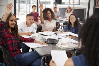 High school kids raise hands, teacher sitting at their desk