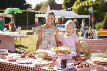 Portrait Of Children Serving On Cake Stall At Busy Summer Garden Fete