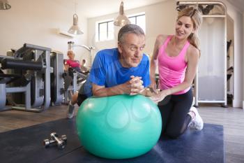 Senior Man Exercising On Swiss Ball Being Encouraged By Personal Trainer In Gym