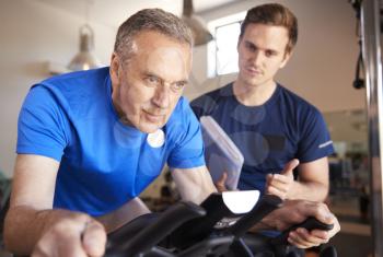 Senior Man Exercising On Cycling Machine Being Encouraged By Personal Trainer In Gym