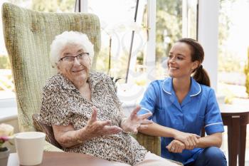 Senior Woman Sitting In Chair And Talking With Nurse In Retirement Home