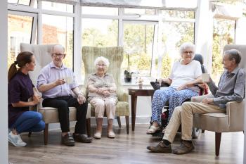 Male And Female Residents Sitting In Chairs And Talking With Carer In Lounge Of Retirement Home