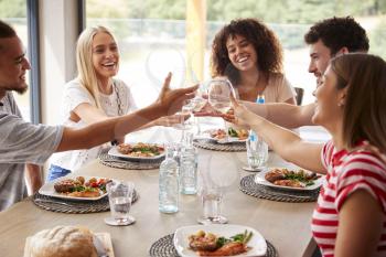 Multi ethnic group of five happy young adult friends laughing and raising glasses to toast during a dinner party