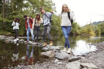 Multi ethnic group of five young adult friends hold hands walking on rocks to cross a stream during a hike