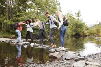 Multi ethnic group of five young adult friends hold hands balancing on rocks to cross a stream during a hike