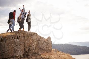 A group of five happy young adult friends cheer with their arms in the air at the summit of a mountain during a hike