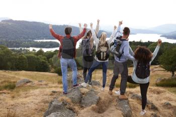 A group of five young adult friends celebrate arriving at the summit after a mountain hike, back view