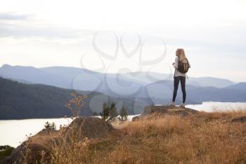 A young adult Caucasian woman standing alone on the rock after hiking, admiring lake view, back view