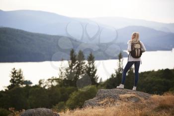 A young adult Caucasian woman standing alone on a peak after hiking, admiring lake view, back view