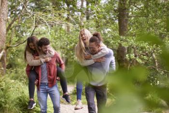 Four smiling young adult friends walking in a forest during a hike, men piggybacking their girlfriends