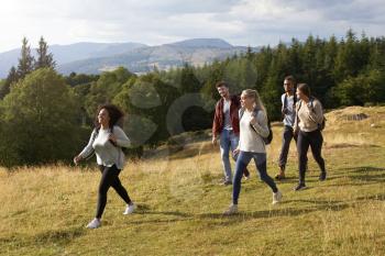 A multi ethnic group of five young adult friends smile while walking on a rural path during a mountain hike, side view