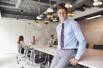 Portrait Of Businessman Standing In Modern Boardroom With Colleagues Meeting Around Table In Background