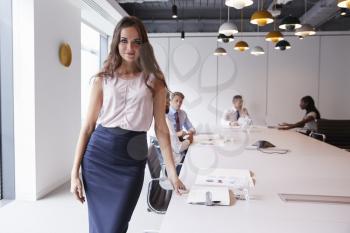 Portrait Of Businesswoman Standing In Modern Boardroom With Colleagues Meeting Around Table In Background