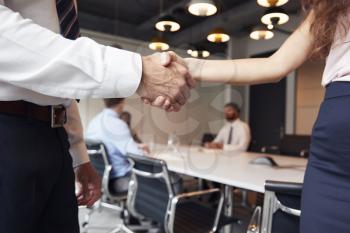 Close Up Of Businessman And Businesswoman Shaking Hands In Modern Boardroom With Colleagues Meeting Around Table In Background