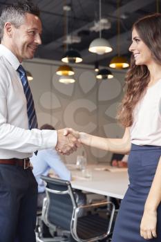 Businessman And Businesswoman Shaking Hands In Modern Boardroom With Colleagues Meeting Around Table In Background