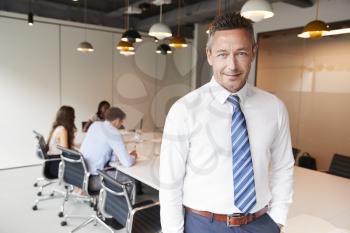 Portrait Of Mature Businessman Standing In Modern Boardroom With Colleagues Meeting Around Table In Background