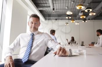 Portrait Of Mature Businessman Sitting In Modern Boardroom With Colleagues Meeting Around Table In Background