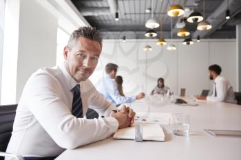 Portrait Of Mature Businessman Sitting In Modern Boardroom With Colleagues Meeting Around Table In Background