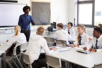 Male High School Teacher Standing Next To Interactive Whiteboard And Teaching Lesson To Pupils Wearing Uniform