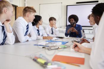 Male High School Teacher Sitting At Table With Teenage Pupils Wearing Uniform Teaching Lesson