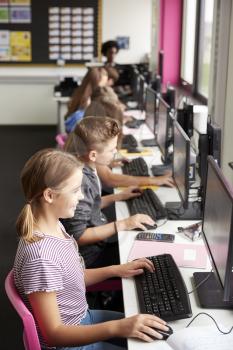 Line Of High School Students Working at Screens In Computer Class With Female Teacher In Background