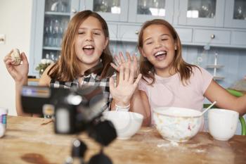 Two pre-teen girls video blogging in the kitchen waving to camera