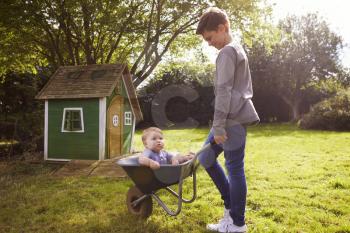 Boy Pushing Baby Brother In Garden Wheelbarrow
