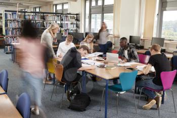 Busy College Library With Teacher Helping Students At Table