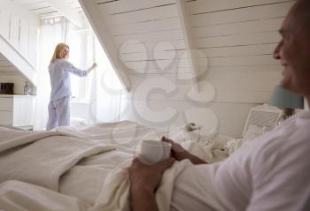 Woman Opens Curtains And Looks Out Of Window As Man Lies In Bed