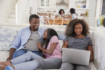 Family With Teenage Daughters Sitting On Sofa With Laptop