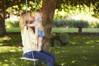 Mother Kissing Baby Son As They Sit On Garden Swing