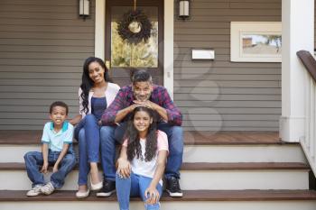 Family With Children Sit On Steps Leading Up To Porch Of Home