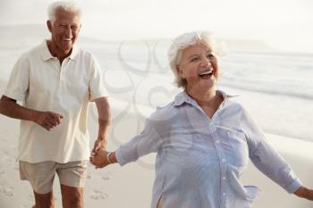 Senior Retired Couple Running Along Beach Hand In Hand Together