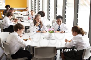 Group Of High School Students Wearing Uniform Sitting Around Table And Eating Lunch In Cafeteria