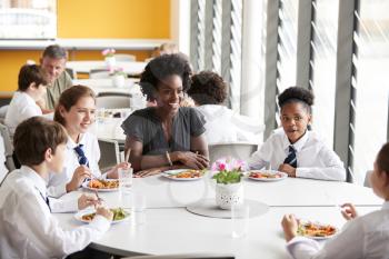 Female Teacher With Group Of High School Students Wearing Uniform Sitting Around Table And Eating Lunch In Cafeteria