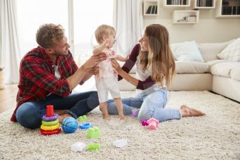 Young couple helping toddler daughter stand in sitting room