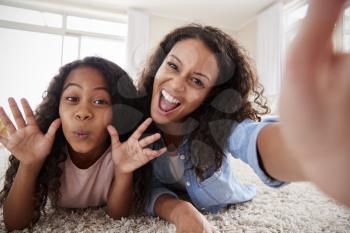 Mother And Daughter Lying On Rug And Posing For Selfie At Home