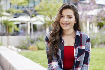 Portrait Of Female High School Student Sitting On Wall Outside College Buildings