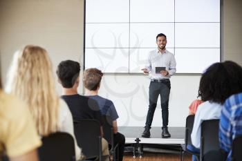 Male Teacher With Digital Tablet Giving Presentation To High School Class In Front Of Screen