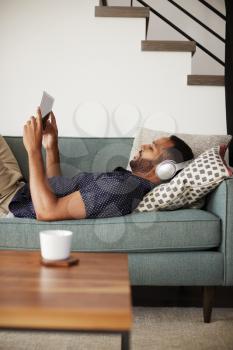 Man Lying On Sofa At Home Wearing Headphones And Watching Movie On Digital Tablet