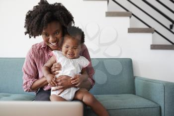 Mother With Baby Daughter Sitting On Sofa At Home Looking At Laptop Computer