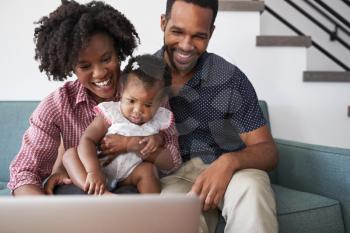 Family With Baby Daughter Sitting On Sofa At Home Looking At Laptop Computer