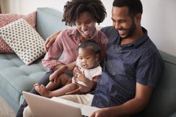 Family With Baby Daughter Sitting On Sofa At Home Looking At Laptop Computer