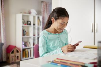 Young Girl Sitting At Desk In Bedroom Using Mobile Phone Whilst Doing Homework