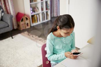 Young Girl Sitting At Desk In Bedroom Using Mobile Phone Whilst Doing Homework