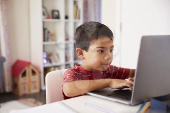 Young Boy Sitting At Desk In Bedroom Using Laptop To Do Homework