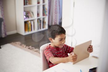 Young Boy Sitting At Desk In Bedroom Using Digital Tablet To Do Homework