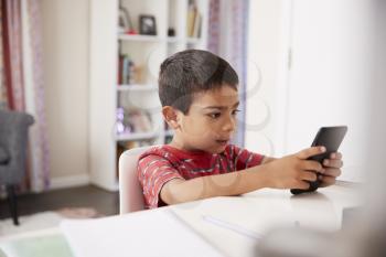 Young Boy Sitting At Desk In Bedroom f Mobile Phone Whilst Doing Homework