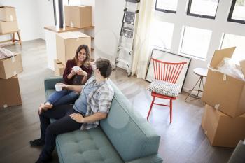 Happy Couple Resting On Sofa Surrounded By Boxes In New Home On Moving Day