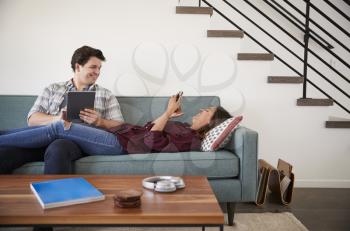 Couple Relaxing On Sofa At Home Using Mobile Phone And Digital Tablet
